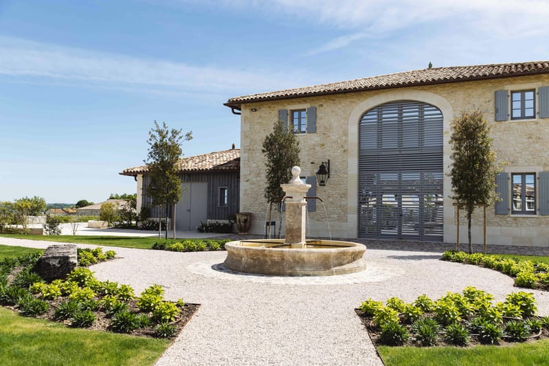 Exterior wide shot of a French country estate featuring a pale limestone facade with terracotta roof tiles, large arched louvered shutters in slate grey, and matching grey window shutters throughout. A circular stone fountain with a central pedestal and running water sits at the center of a gravel courtyard, flanked by neatly landscaped beds with low green shrubs and slender young trees. A secondary outbuilding with grey vertical cladding is visible to the left. No people are present in the image. Potential venue feature image.