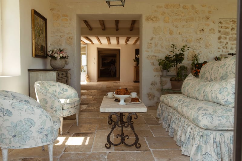 An interior sitting room of a French country property, styled in a classic Provençal manner with no people present. The space features matching floral-upholstered armchairs and a skirted sofa in a soft blue and white botanical print, arranged around a wrought-iron coffee table with a stone top holding white porcelain espresso cups and a plate of croissants. A distressed pale blue-grey sideboard sits against the wall beneath a gold-framed oil painting, topped with a round stone vase holding a loose arrangement of blush pink and white blooms including roses and peonies. Potted citrus trees with visible orange fruit are visible to the right, alongside additional green potted plants. The hallway beyond the arched doorway reveals exposed wooden ceiling beams and stone tile flooring throughout, reinforcing the rustic French farmhouse aesthetic. Wide-angle interior shot taken from the doorway. Potential venue feature image.