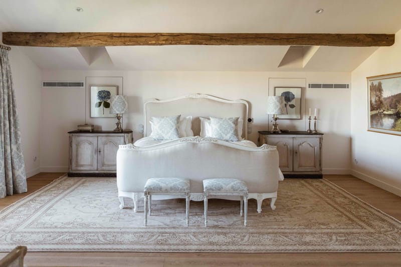 Interior shot of a bridal suite or guest bedroom at a French wedding venue, shown with no people present. The room features a white upholstered French Louis-style bed with an ornate carved headboard and footboard, dressed in white and pale blue damask cushions. Two matching white-painted Louis-style footstools with pale blue damask upholstery are placed at the foot of the bed. Flanking the bed are two distressed grey-painted antique-style bedside cabinets, each topped with a silver candlestick lamp with a white damask shade. Botanical hydrangea prints in matching frames hang symmetrically above each cabinet, and silver candlestick holders are placed on the right cabinet. The room has exposed dark oak ceiling beams, a large neutral-toned patterned area rug over light wood flooring, blue-and-white toile curtains, and cream walls, creating a classic French country aesthetic. Wide shot taken from a straight-on, centred angle. Potential venue feature image.