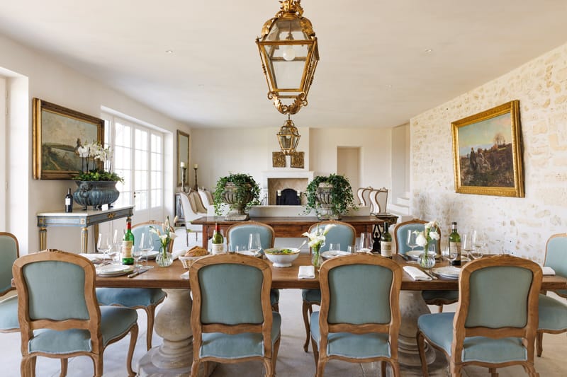 An indoor dining room set for a formal meal, likely a wedding reception dinner or rehearsal dinner, inside what appears to be a French château or manor house. A long rectangular wooden dining table seats approximately twelve, surrounded by Louis XV-style carved oak chairs upholstered in pale blue-grey fabric. The table is set with white plates, crystal wine glasses, white linen napkins, wine bottles, a large white ceramic salad bowl, and small bud vases holding white calla lilies. Two ornate gilded lantern pendants hang from the cream-painted ceiling above the table, and the room features exposed limestone walls, gold-framed oil paintings, a white stone fireplace with herringbone tile surround, and stone urn planters filled with trailing ivy. The overall decor palette is cream, warm oak, pale blue, and gold, with a classic French provincial style. Wide establishing shot taken from one end of the table. Potential venue feature image.