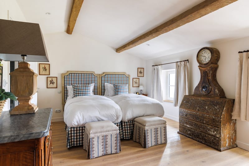 Interior shot of a guest bedroom within what appears to be a French country property. The room features two single beds with blue and white gingham upholstered headboards and matching bed skirts, dressed in white bedding with coordinating checked cushions. Two small upholstered ottomans in a blue and brown geometric stripe pattern sit at the foot of each bed. The room is styled in a classic French country aesthetic with exposed wooden ceiling beams, light oak flooring, and white walls. A large antique Mora-style grandfather clock with heavily distressed dark wood finish stands against the right wall alongside a matching antique bureau. Framed equestrian prints in red-orange mounts are hung on the walls, and a substantial wooden table lamp with a taupe shade sits on a dark marble-topped chest to the left. Natural linen curtains frame a window to the right. Wide-angle interior shot. Potential venue feature image.