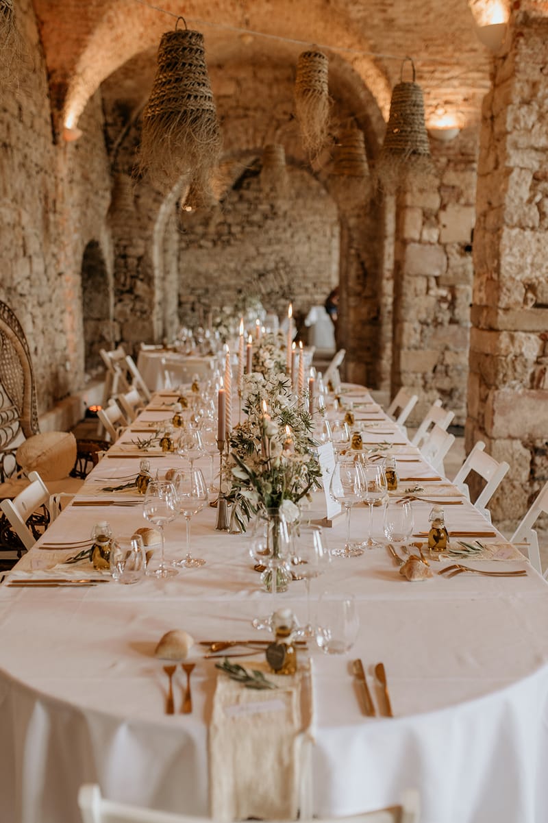 Vaulted stone reception with rattan pendants blush taper candles white floral bud vases olive oil favors and peacock chair