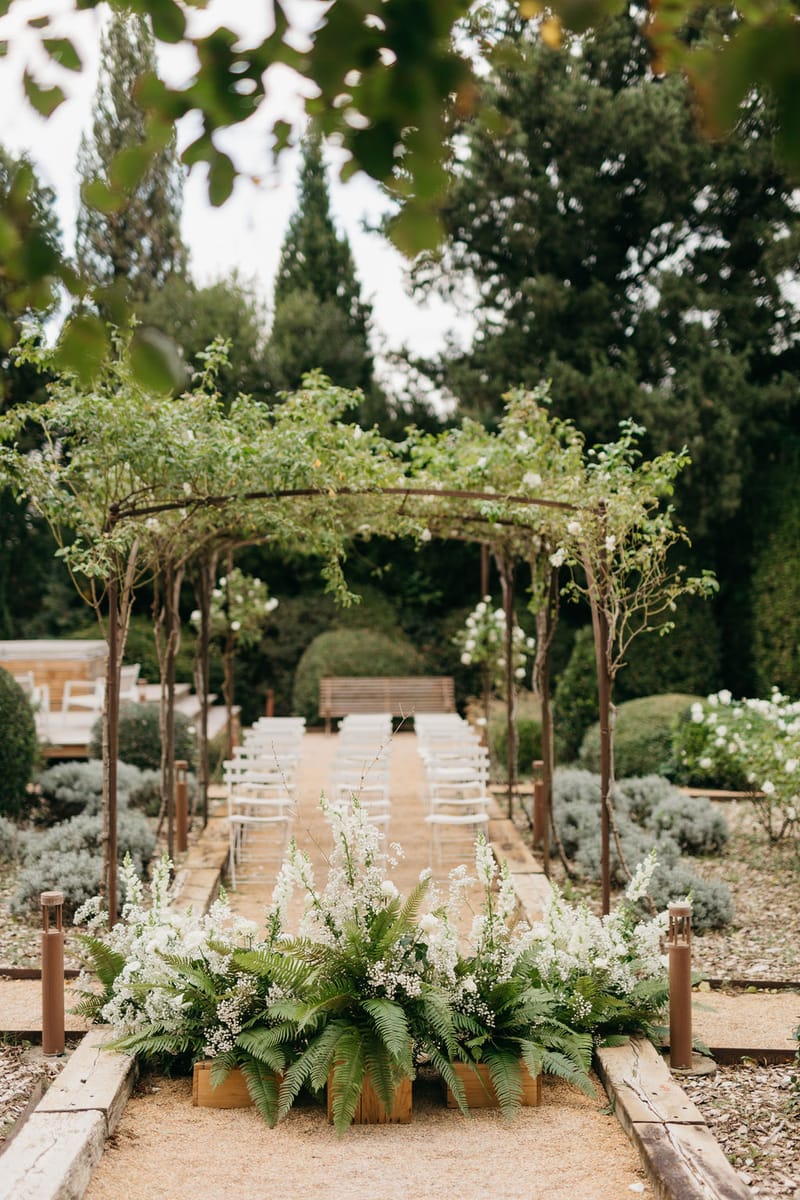 Ceremony aisle under iron pergola with climbing roses white chairs fern arrangements and box hedging