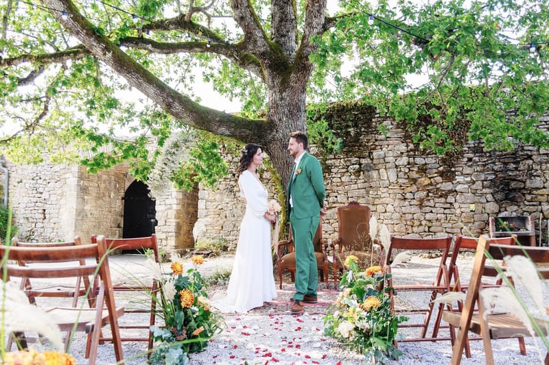 Couple at outdoor ceremony beneath circular floral arch in ruined stone courtyard with boho dahlia aisle decor
