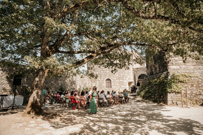 An outdoor wedding ceremony taking place in a courtyard enclosed by old stone walls, likely a historic château or fortified site in France. Approximately 40–50 guests are seated on wooden folding chairs arranged in rows, facing toward a small elevated area at the far end where two figures — likely the officiant and a seated person — are positioned near a stone archway. The guests are dressed in colorful summer attire including coral red, sage green, and teal dresses, giving the gathering a relaxed, warm-weather feel. A large mature tree dominates the foreground, casting dappled shade across the courtyard floor, and a DJ or sound setup with a speaker stand is visible to the left. The shot is a wide-angle view taken from behind the guests, framing the ceremony within the tree canopy and stone architecture. Potential venue feature image.