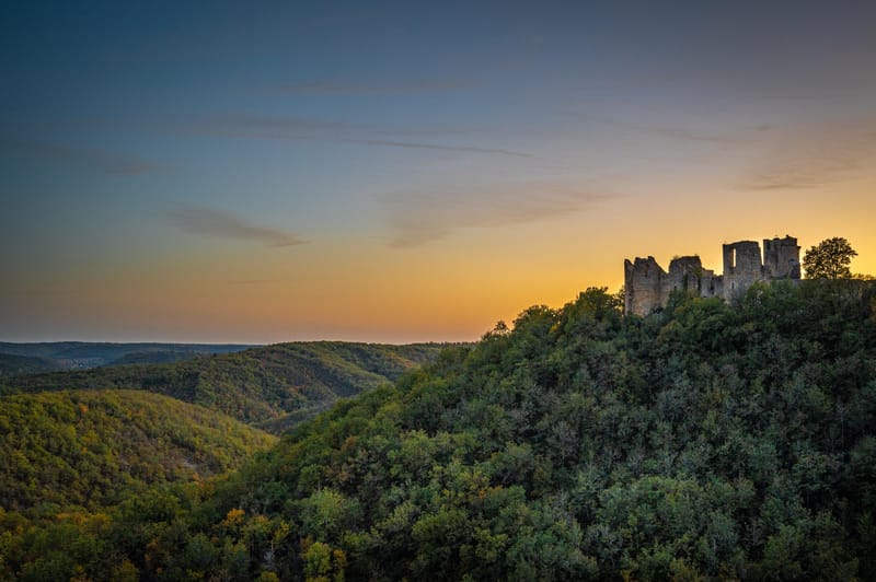 Wide-angle aerial or elevated landscape shot showing the ruins of a medieval stone castle or fortification perched on a densely forested hilltop, with rolling wooded valleys extending into the distance. No people, wedding party, or wedding-related decor are visible in this image — it appears to be a standalone venue or location establishing shot taken at dusk, with a warm amber and golden horizon transitioning to a blue-grey upper sky. The castle ruins feature multiple remaining stone towers and walls in partial collapse. Potential venue feature image.