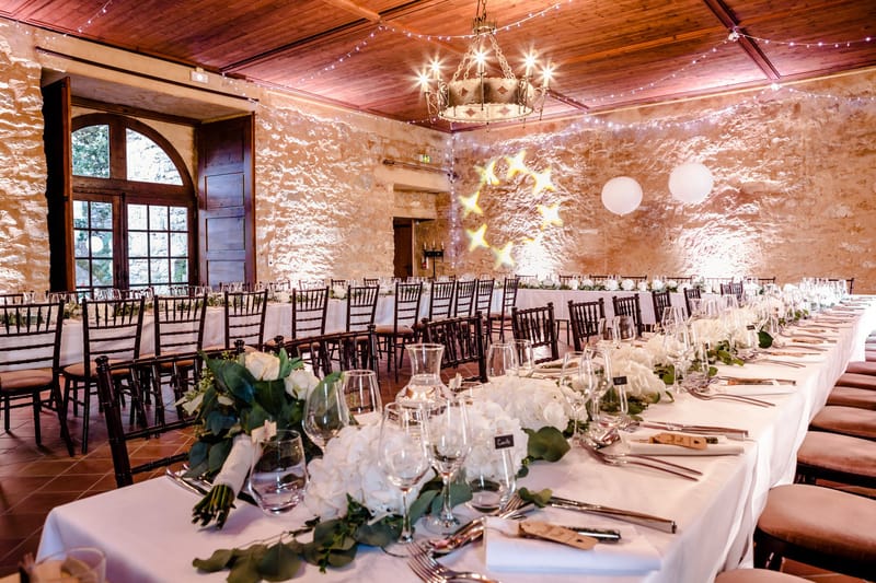 A wedding reception room set for dinner inside a rustic stone-walled venue with a warm wood-paneled ceiling. The space features multiple long banquet tables dressed in white linen, lined with dark wood Chiavari chairs with tan cushions. Table centerpieces consist of white roses and hydrangeas with trailing eucalyptus garlands running the length of each table, accompanied by crystal glassware, silver cutlery, white folded napkins, and small chalkboard place cards. Lighting includes a wrought-iron chandelier overhead, strings of fairy lights crisscrossing the ceiling, warm uplighting illuminating the stone walls, and two large white paper lantern globes mounted on the back wall. A tall arched wooden door with a grid-pane window is visible on the left wall. The overall decor palette is white and green with warm amber lighting, giving the room a classic rustic feel. Wide-angle interior shot with no people present. Potential venue feature image.
