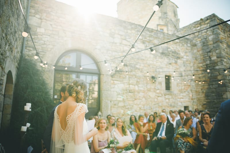 An outdoor wedding ceremony taking place in the courtyard of a stone château or castle, with the couple standing at the altar facing approximately 40–50 seated guests. The bride is seen from behind, wearing a low-back ivory gown with intricate crochet or cutwork lace detailing on the back and a flowing veil; her hair is styled in an updo with small floral pins. The groom stands beside her in a dark suit. String lights with globe bulbs are strung across the courtyard overhead, and pillar candles on tall stands are visible to the left. Guests are dressed in a mix of summer attire including dusty mauve, mustard yellow, and navy outfits, and at least one guest holds a white and greenery bouquet. The image is a wide shot taken from behind the couple, with strong sun flare creating warm, hazy light across the scene. Potential venue feature image.