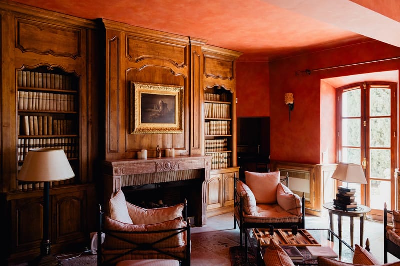 Chateau library with terracotta walls, floor-to-ceiling bookshelves, stone fireplace, and gingham armchairs