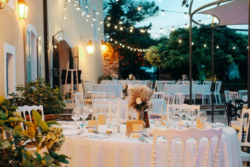 An outdoor wedding reception setup photographed at dusk in the courtyard of a French manor or bastide, with warm globe string lights strung overhead and vintage-style wall lanterns illuminating the space. Multiple round tables are dressed in white linen tablecloths and surrounded by white Napoleon chairs, set for a large number of guests across the courtyard. The foreground table features a centerpiece of pampas grass and a single cream rose in a glass vase, flanked by taper candles, wine glasses, kraft paper place cards, and small terracotta-toned favor boxes. The decor palette is neutral and boho-rustic, combining white, cream, and warm tan tones. This is a wide-angle shot taken at table level, capturing both the detailed foreground place setting and the full reception layout in the background. Potential venue feature image.