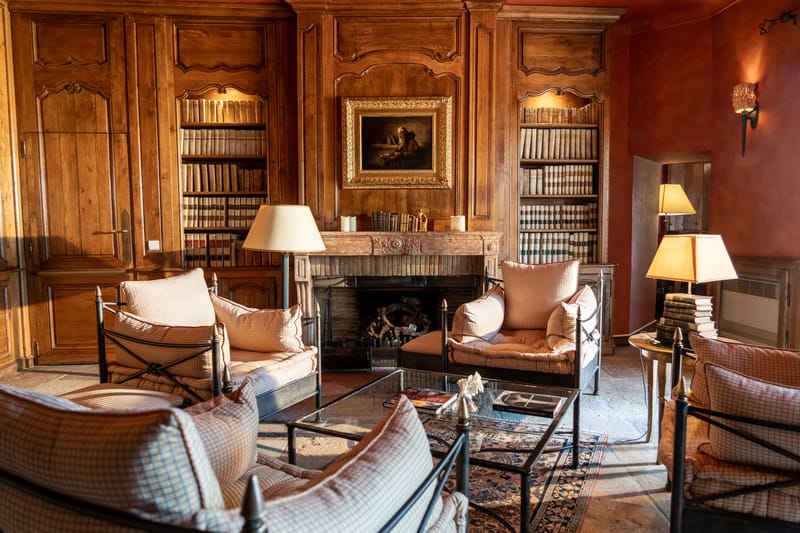 Chateau library with carved wood paneling, antique books, stone fireplace, and checked armchairs on Persian rug