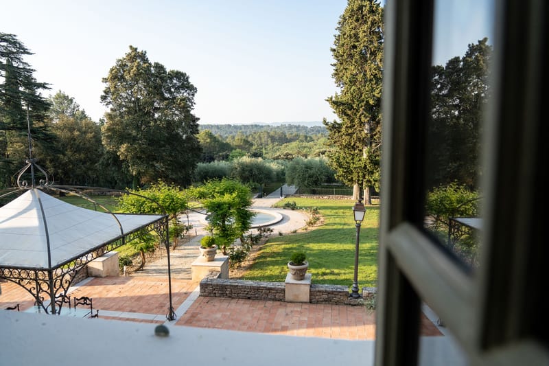 Elevated view of French domaine grounds with wrought-iron pergola, gravel driveway, manicured lawn, and mature trees