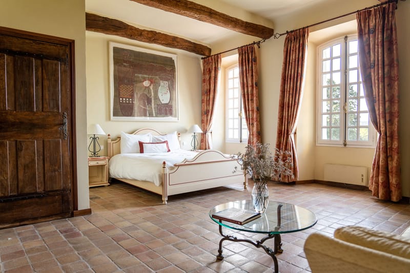 Interior wide shot of a guest bedroom in what appears to be a French chateau or mas, featuring terracotta tile flooring, exposed dark wood ceiling beams, and cream-painted walls. The room contains a white-painted wooden double bed with red trim detail, dressed in white bedding with burgundy accent cushions, flanked by cream bedside tables with iron lyre-shaped bases and small white lampshades. Two arched French windows are dressed with coral and dusty rose floral-print curtains, and a round wrought-iron glass-top coffee table in the foreground holds a glass vase of dried pale flowers and a hardcover book. A large framed artwork in muted tones hangs above the bed, and a heavy dark wood door is visible on the left. The overall decor palette is warm terracotta, cream, and dusty rose with a classic Provençal style. Potential venue feature image.