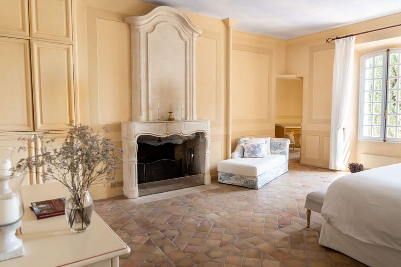 A wide interior shot of a spacious château bedroom suite, likely used as a bridal getting-ready space. The room features warm butter-yellow panelled walls, terracotta diamond-pattern floor tiles, and a large carved limestone fireplace with an ornate arched overmantel. Two white taper candles and a small amber glass object sit on the mantelpiece. A blue and white toile de Jouy daybed with matching cushions is positioned in the center of the room, and a white-linened bed is partially visible at the right edge of the frame. A white side table in the foreground holds a glass vase with dried lavender-grey botanicals and a stack of books. Sheer white curtains frame tall French windows letting in natural light. The overall décor palette is soft yellow, white, and muted blue with classic French provincial styling. Potential venue feature image.