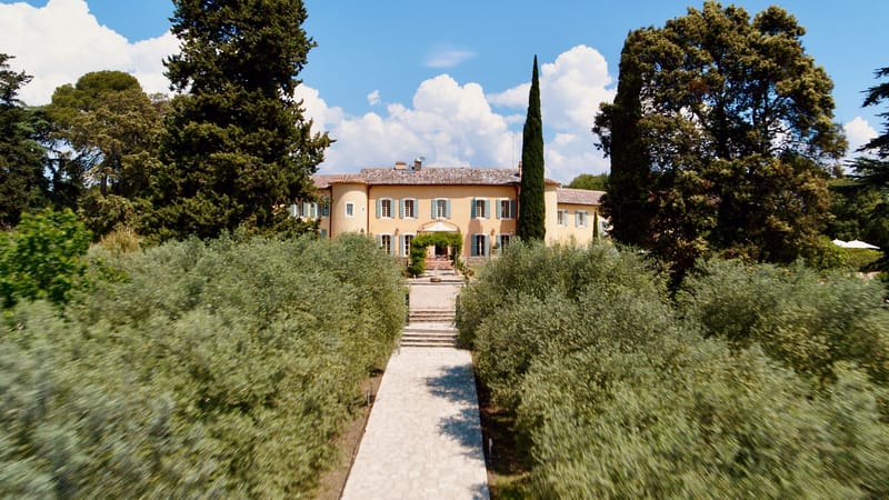 Aerial view of ochre Provencal bastide with blue shutters, olive tree allee, and cypress trees along stone pathway