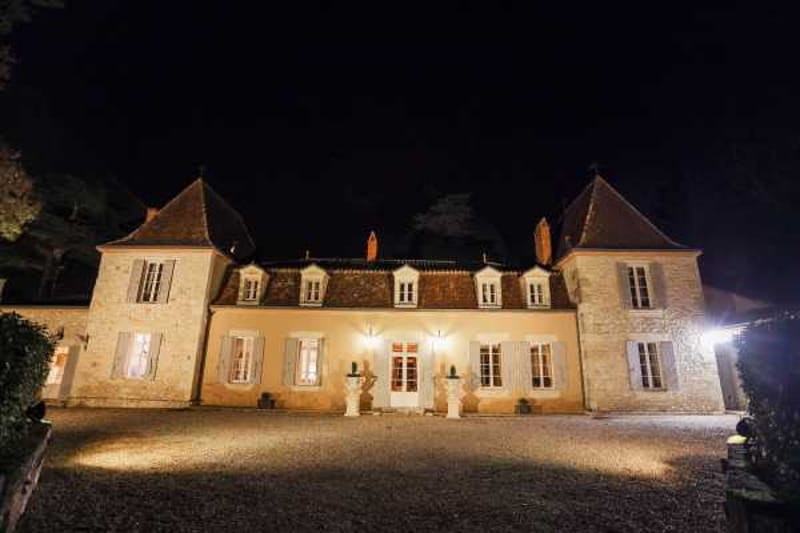A wide-angle exterior night shot of a French château with classic stone architecture, featuring two flanking tower wings with steeply pitched tile roofs and white shuttered windows. The building is illuminated by warm golden uplighting that highlights the pale stone facade, with interior lights glowing through the ground-floor French doors and windows. The entrance is framed by two decorative stone urns or pedestals flanking the central doorway. A gravel forecourt extends in the foreground, reflecting the warm light from the building. No people are visible in the frame. Potential venue feature image.