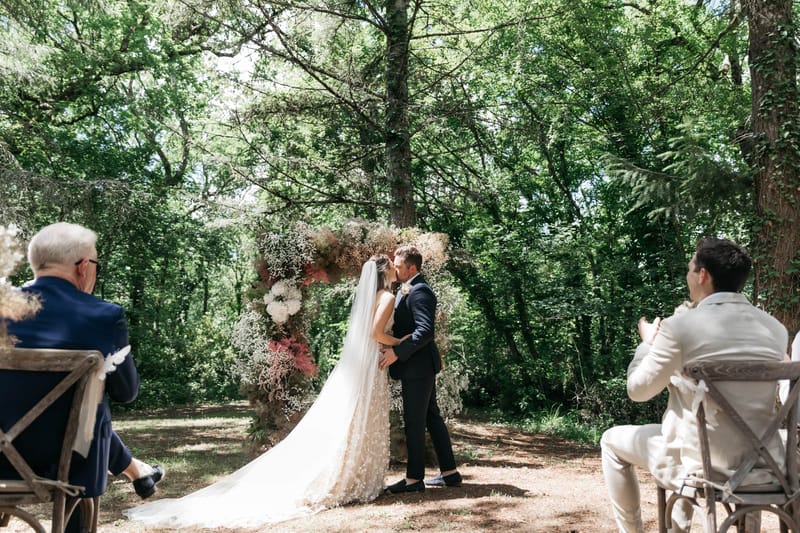 First kiss under circular floral arch in woodland clearing with cross-back chairs and guests