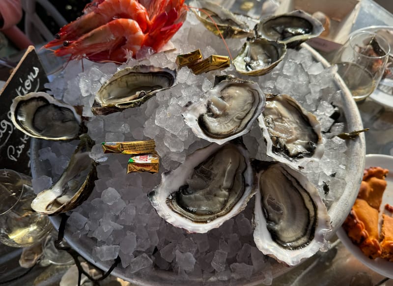 This image does not appear to be a wedding photograph. It shows a close-up overhead shot of a seafood platter at what looks like a casual outdoor dining setting, featuring freshly shucked oysters and whole prawns arranged on crushed ice on a round metal tray, with small individual Président butter packets scattered among the shells. Two glasses of white wine are partially visible at the edges, along with what appears to be a chalkboard menu sign and a partial view of a crab dish. This image is not suitable for a wedding photography content library.