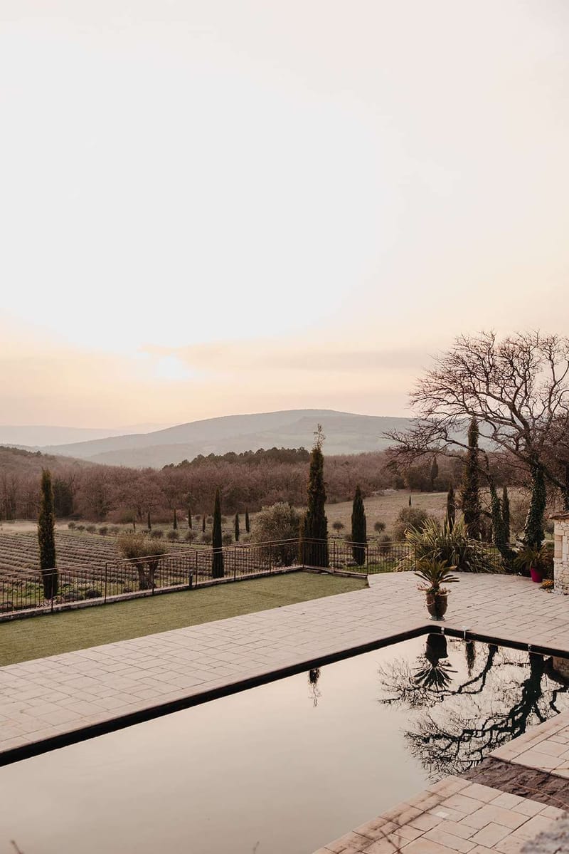 A wide exterior shot of a Provençal estate venue at dusk, with no people or wedding activity visible. The foreground features a dark-edged infinity or reflecting pool set into a pale stone terrace, with the still water mirroring surrounding trees. A manicured lawn area and a potted palm sit beside the pool, bordered by a wrought-iron railing. Beyond the grounds, rows of what appear to be lavender fields and cypress trees line the property, with rolling hills and a warm peach-toned horizon in the background. The overall palette is muted , warm stone, deep green cypress, and soft amber light. Potential venue feature image.