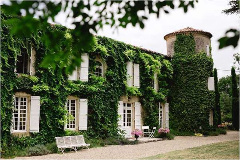 Ivy-covered French manor house with white shutters, round tower, gravel terrace, and wrought-iron bench