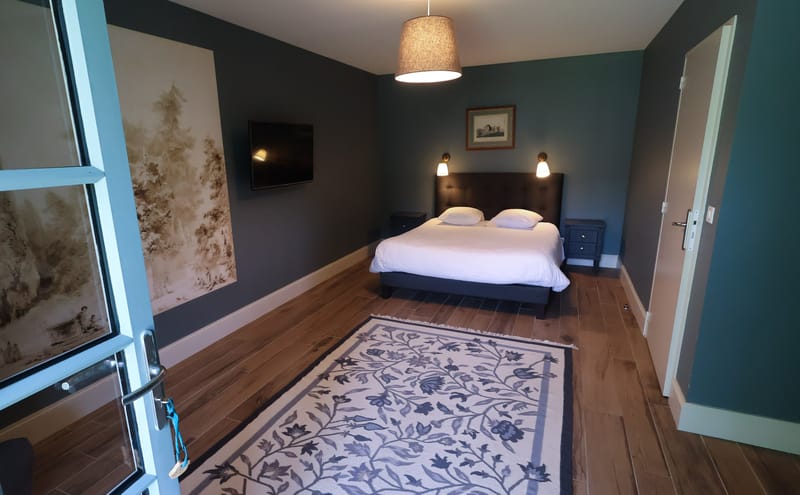 Interior wide shot of a guest bedroom, likely part of a wedding venue's on-site accommodation. The room features dark teal-painted walls, warm wood-tone flooring, and a double bed with a dark upholstered headboard and white bedding, flanked by two navy blue bedside tables with small wall-mounted sconce lights. A large floral-patterned rug in cream and blue-grey tones covers the floor in the foreground. One wall features a decorative mural or wallpaper panel with a sepia-toned landscape motif, and a flat-screen TV is mounted above it. A linen pendant light hangs from the ceiling, and a framed print hangs above the bed. The room is viewed from the doorway entrance, with a glazed door visible on the left. Potential venue feature image.