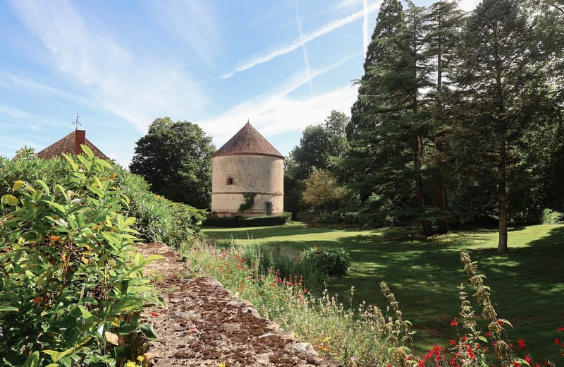 French country estate grounds with circular stone dovecote, terracotta roof, open lawn, and wildflower border