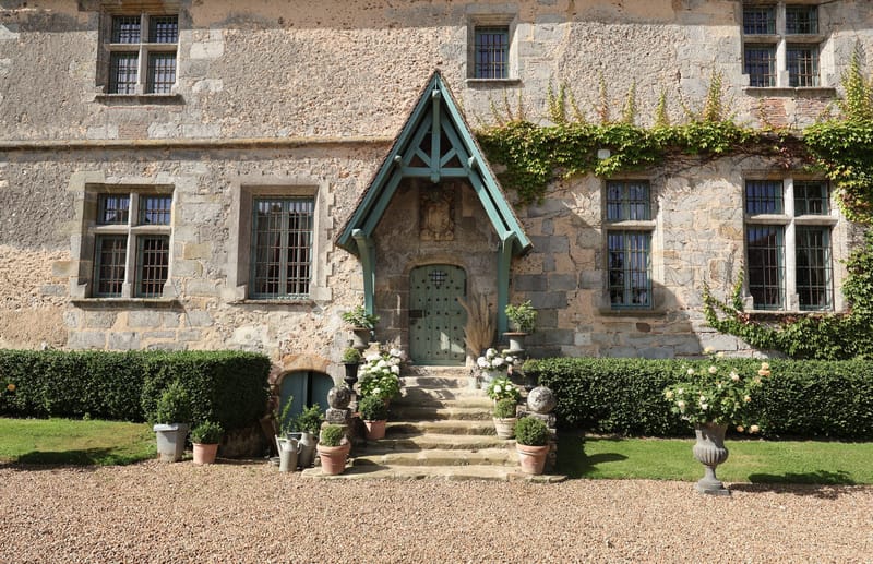 French chateau entrance with teal-painted door and porch canopy, flanked by topiary and hydrangeas on gravel path