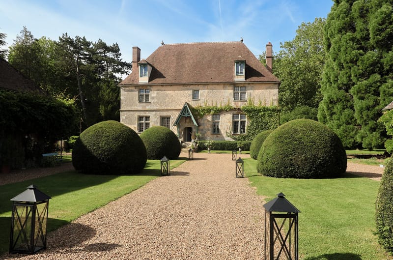 Stone manor with mansard roof and green entrance porch approached by topiary-lined driveway with lanterns