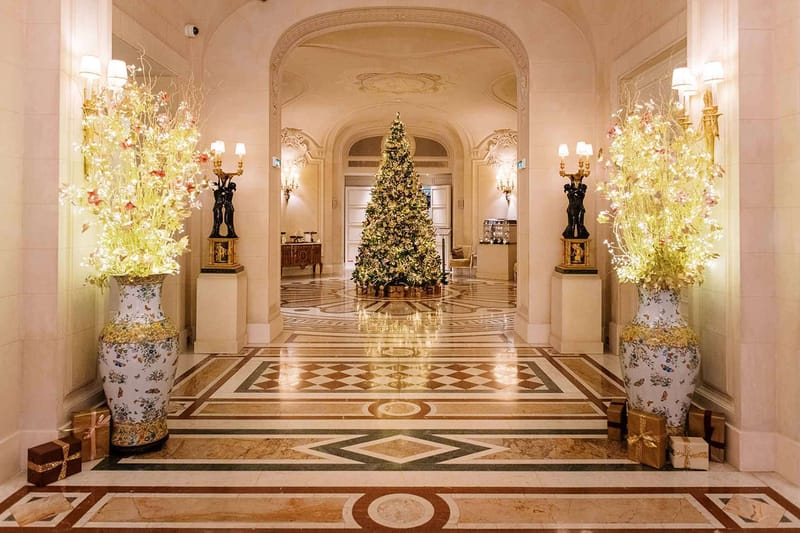 A wide shot of an indoor grand hotel or palace lobby decorated for Christmas, with no people present. The hallway features an ornate inlaid marble floor in cream, green, and brown geometric patterns, arched doorways with decorative plasterwork ceilings, and warm wall sconce lighting. Two large decorative porcelain vases with floral botanical motifs flank the entrance, each filled with tall arrangements of chartreuse and pale pink branches with fairy lights woven through. A fully decorated Christmas tree with warm white lights and gold ornaments stands at the far end of the corridor, surrounded by wrapped gifts in gold and brown at its base. Additional wrapped gifts in pink and gold are placed near the foreground vases, and pairs of bronze figural candelabras on stone pedestals line the corridor. The overall decor palette is gold, cream, and warm white, with a classic French palatial interior style. Potential venue feature image.