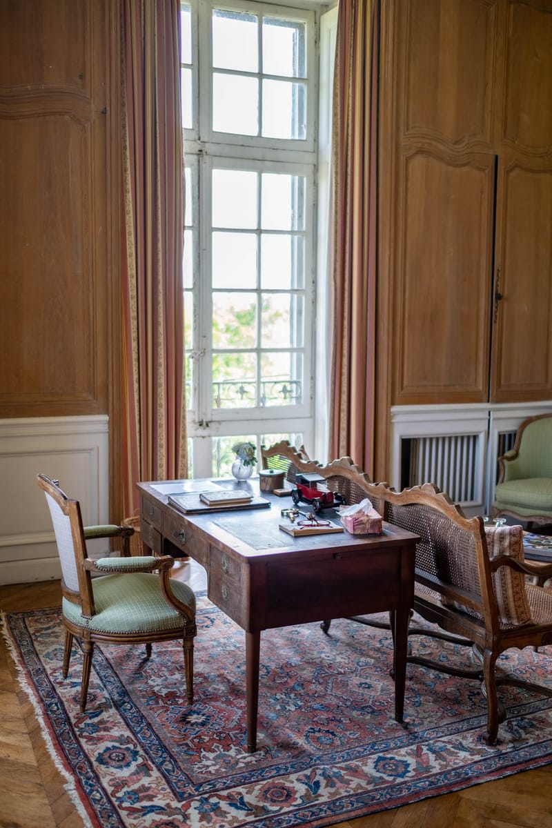 Interior room shot of a château study or salon, with no people present. The room features floor-to-ceiling warm honey-toned wood panelling, a tall multi-pane white-framed French window with salmon and gold striped curtains, and a herringbone parquet floor. A dark mahogany writing desk sits at the centre on a large Persian-style rug in dusty rose, navy blue, and ivory tones. Around the desk are two Louis XVI-style armchairs with sage green upholstered seats and a cane-backed settee. The desk surface holds personal items including books, a small red toy car, a small white vase with greenery, and a gift box. A cast-iron radiator is visible in the background alongside a partially visible green sofa. The composition is a wide interior shot taken from a slightly elevated angle. Potential venue feature image.