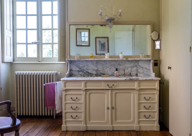 Interior shot of a château bathroom, likely part of a bridal suite or getting-ready space. The room features a large antique cream-painted double vanity with a grey-veined white marble countertop and backsplash, silver cross-handle faucets, and multiple drawers with silver bail pulls. A large antique mirror with aged foxing sits above the vanity, reflecting a Murano-style glass chandelier and a framed artwork on the opposite wall. The room has wide-plank wood flooring, a white cast-iron radiator, tall white-framed windows letting in natural light, and a monogrammed white towel alongside a pink towel draped over the radiator. A vintage wooden chair is partially visible in the lower left corner. Wide shot, no people present. Potential venue feature image.