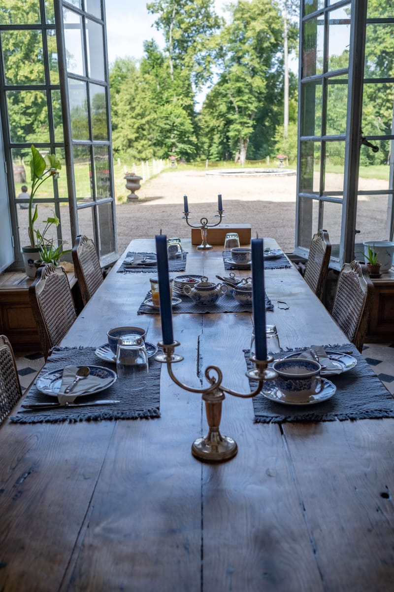 A styled interior dining room shot showing a large rustic oak farmhouse table set for approximately six guests, photographed from one end with a straight-on perspective. The table is dressed with grey woven placemats with fringe edges, blue-and-white porcelain teacups and saucers, white plates with navy blue napkins, glassware, and silver candelabras holding tall navy blue taper candles. A blue-and-white ceramic serving bowl sits at the center of the table. The chairs are wicker-backed wooden dining chairs. The room opens through tall white-framed French doors onto a gravel courtyard with stone urns and formal garden allées beyond, suggesting a classic French château or manor house setting. The overall decor palette is navy blue, grey, and white with warm wood tones, consistent with a classic French country styling. Potential venue feature image.