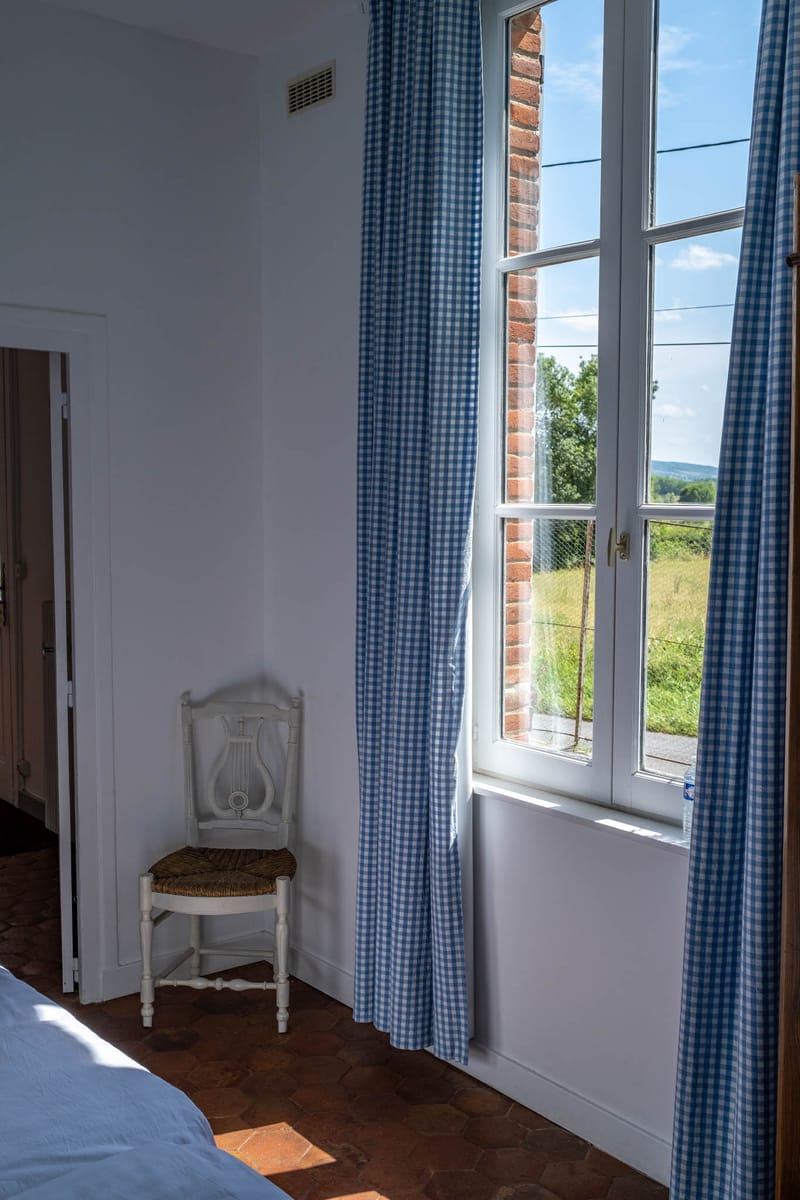 French country bedroom with white antique lyre-back chair, blue gingham curtains, and terracotta floor