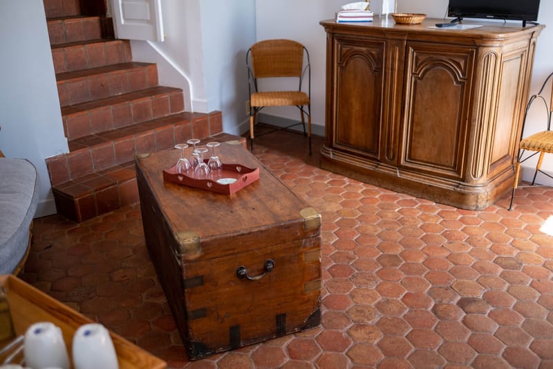 Rustic French country living room with terracotta floor tiles, wooden trunk, carved armoire, and brick staircase