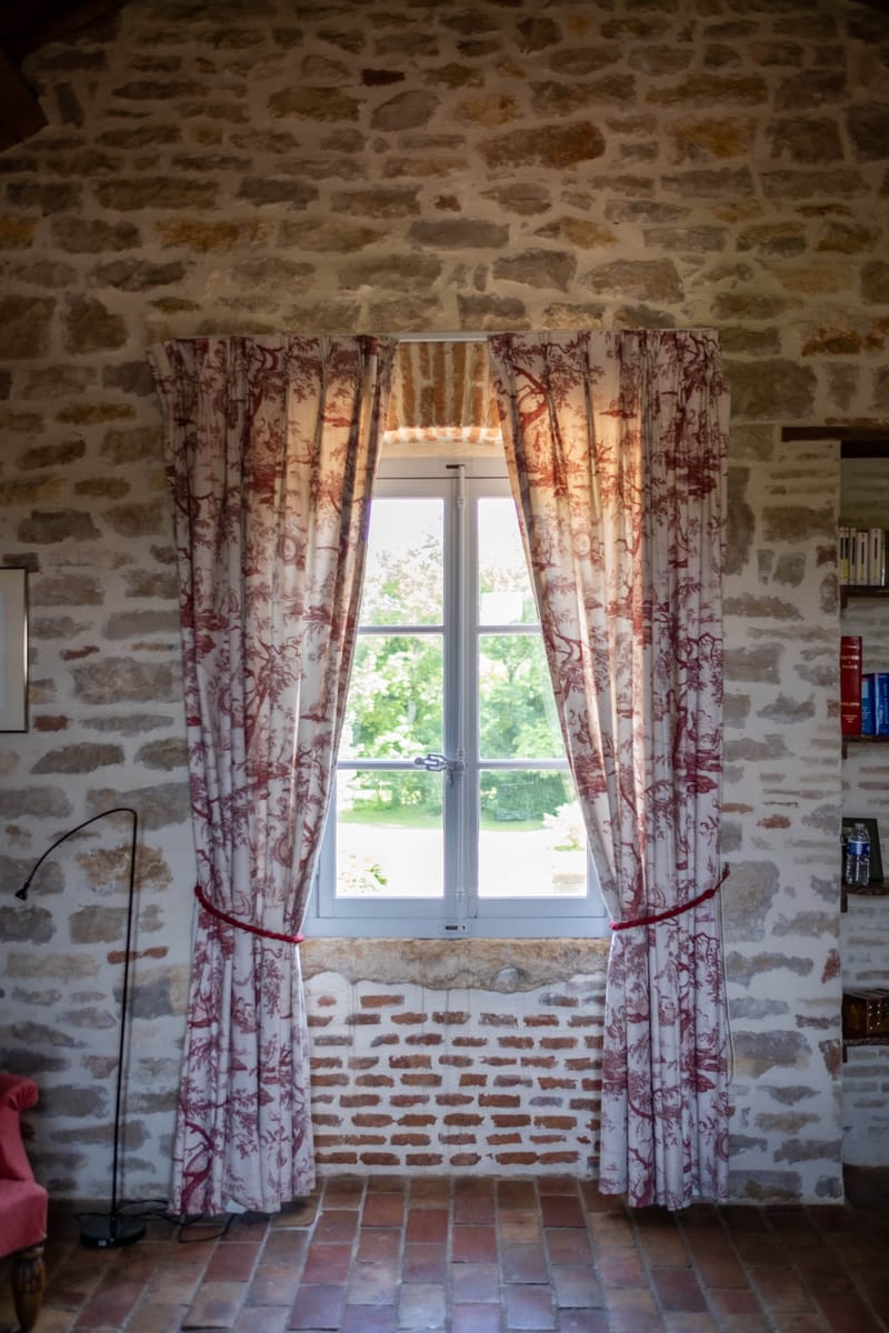 French stone room interior with toile de Jouy curtains, terracotta floor tiles, and casement window overlooking grounds