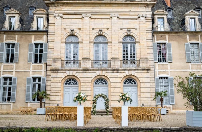 Outdoor ceremony setup with cross-back chairs and circular floral arch before chateau facade
