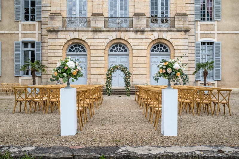 Outdoor ceremony with cross-back chairs, hydrangea urns, and floral arch at chateau entrance