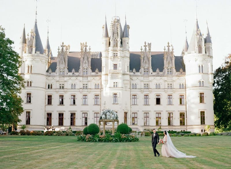 Bride in blush ballgown and groom in tuxedo posing on lawn before a white Gothic-Renaissance chateau with turrets