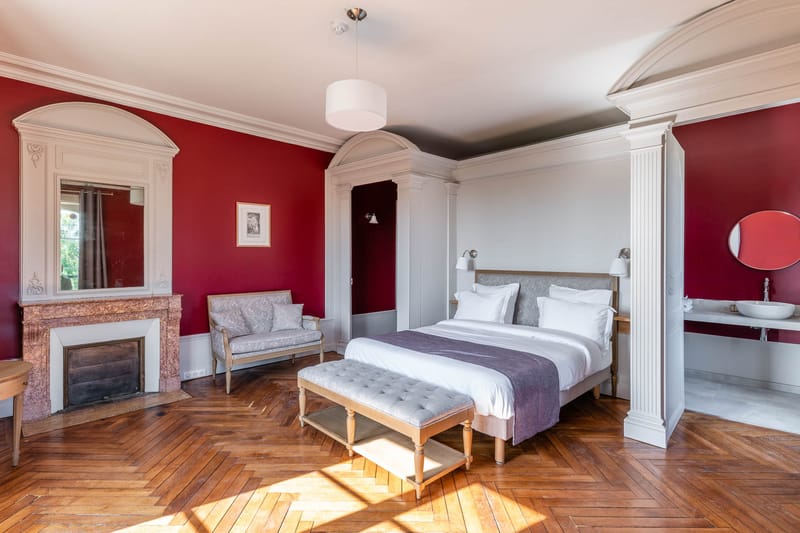 Chateau guest bedroom with crimson walls, white plasterwork, marble fireplace, herringbone parquet floor, and grey furnish...