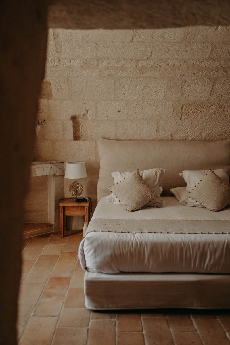 Interior shot of a guest bedroom within what appears to be a French stone-walled property, photographed through a doorway or archway opening. The room features a low-profile double bed dressed in cream and warm sand-toned linen, with textured cushions trimmed with small pom-pom details. A small wooden bedside table holds a white table lamp and a gold dried palm leaf decorative piece. The flooring is terracotta tile, and the walls are exposed cut limestone blocks. The overall decor palette is neutral — cream, sand, and warm terracotta — with a rustic-meets-minimal styling approach. Wide interior shot framed by the doorway edge. Potential venue feature image.