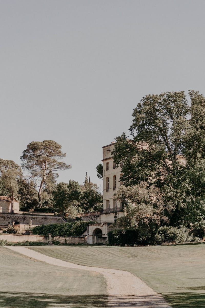 Cream stone chateau viewed across manicured lawn with gravel path, balustrades, and mature trees