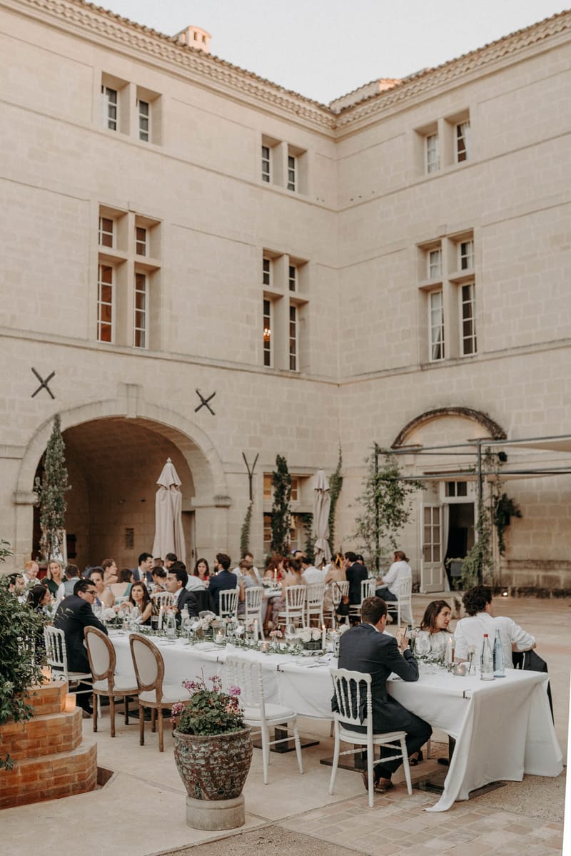 An outdoor wedding reception dinner is taking place in the interior courtyard of a large French château, with approximately 50–60 guests seated at long rectangular tables covered in white linen. The tables are set with white Chiavari and Louis XVI-style chairs, glassware, candles, and low floral centerpieces featuring light pink and white blooms with greenery. The décor palette is classic and understated, with white linens, candlelight, and trailing greenery adding softness to the stone surroundings. A large aged terracotta pot planted with pink cyclamen and trailing foliage anchors the foreground, and climbing plants frame the arched passageways of the château's façade. The image is a wide shot taken from a slightly elevated angle, capturing both the dinner scene and the full three-story limestone building behind it. Potential venue feature image.