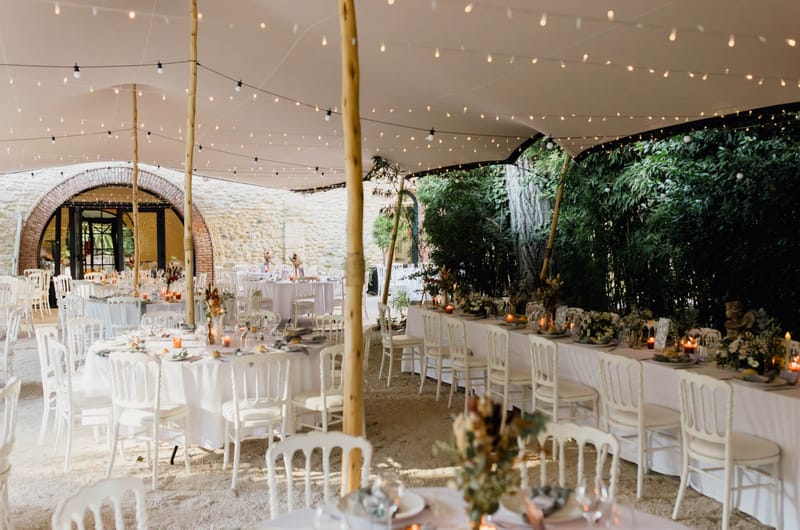 A wide shot of a wedding reception setup inside a stretch tent (tente de réception) erected in what appears to be a courtyard or outdoor space alongside a stone and brick wall featuring arched doorways. The tent is supported by natural wood poles and strung with warm-toned festoon globe lights across the ceiling. The layout combines round tables with white linen and white Napoleon chairs on the left side, and a long rectangular banquet table with white linen running along the right side near a backdrop of dense green bamboo. Centerpieces feature low floral arrangements in warm amber, rust, and terracotta tones with dried and fresh botanicals, accompanied by clusters of amber-glowing votive candles. Place settings include glassware, plates, and folded napkins throughout. The overall decor palette is white, warm gold, and terracotta, with a relaxed rustic-natural styling aesthetic. Potential venue feature image.
