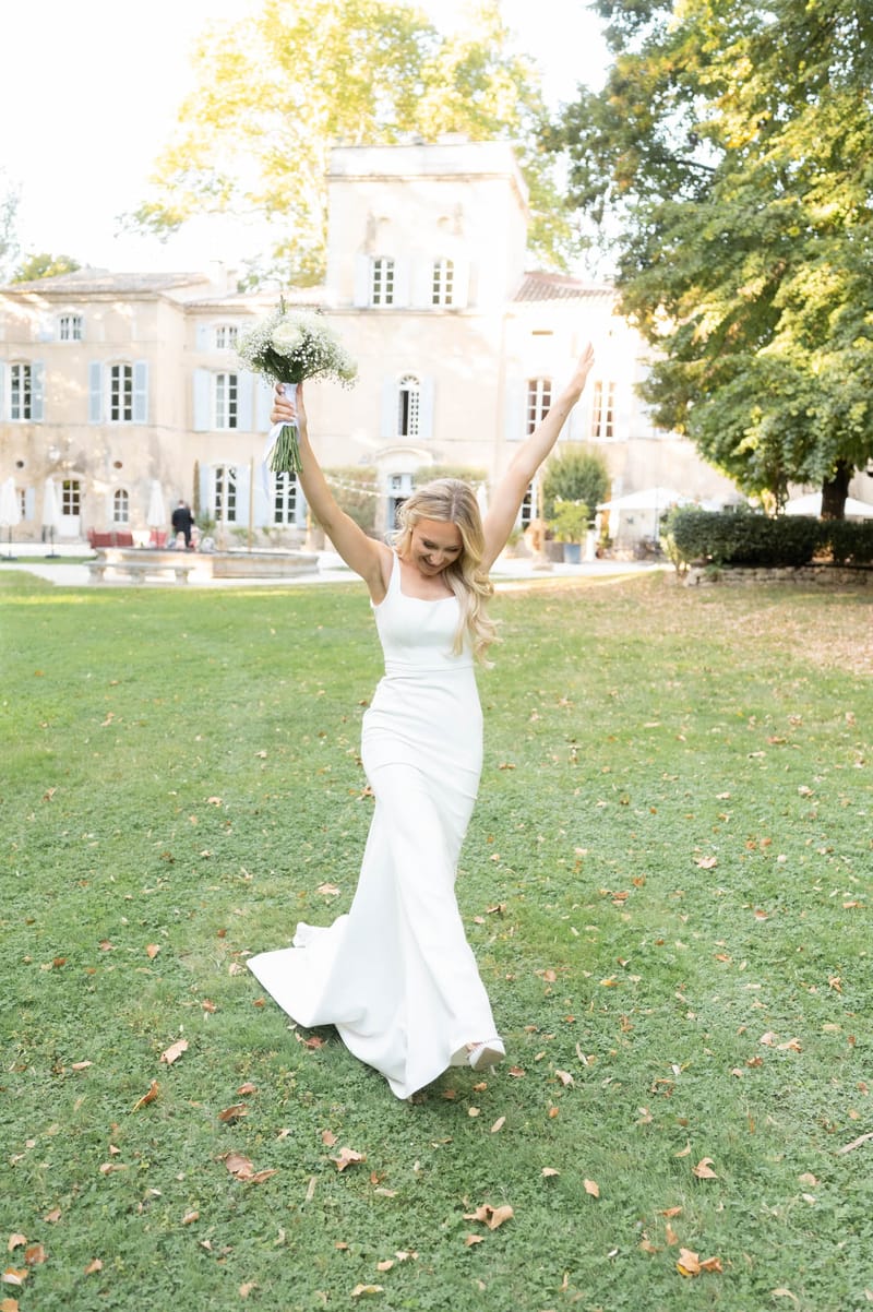 Bride laughing with bouquet raised on chateau lawn wearing ivory satin gown with Provencal manor behind