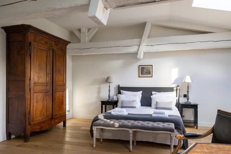 Interior shot of a guest bedroom, likely within a French château or manor house venue. The room features exposed white-painted timber ceiling beams, light oak hardwood flooring, and white walls. The bed has a dark charcoal iron frame dressed in white and grey linen with folded towels placed on top, flanked by two dark bedside tables with ivory lamp shades. A large antique French armoire in warm walnut wood stands to the left, and a grey tufted upholstered bench sits at the foot of the bed. A leather chair is partially visible in the foreground right. The styling mixes antique French provincial furniture with a clean, neutral grey and white palette. Wide interior shot. Potential venue feature image.