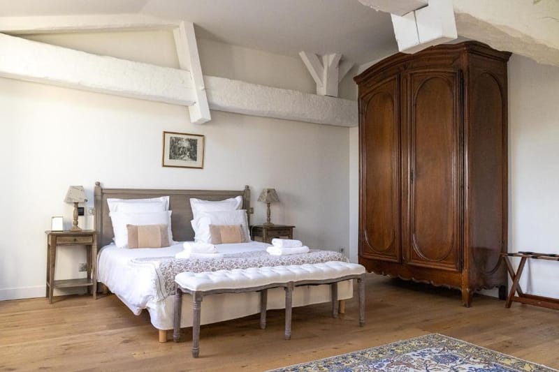 This image shows a guest bedroom interior rather than a wedding photograph. The room features a double bed with a grey-toned wooden headboard dressed in white linens and blush-toned accent pillows, flanked by two rustic wooden nightstands with small lamp shades. A large antique French armoire in dark walnut wood stands prominently to the right. The room has exposed white-painted ceiling beams, light wood plank flooring, a tufted upholstered bench at the foot of the bed, and a patterned area rug in the foreground. The overall styling is classic French country with a neutral white and warm wood palette. Potential venue feature image.