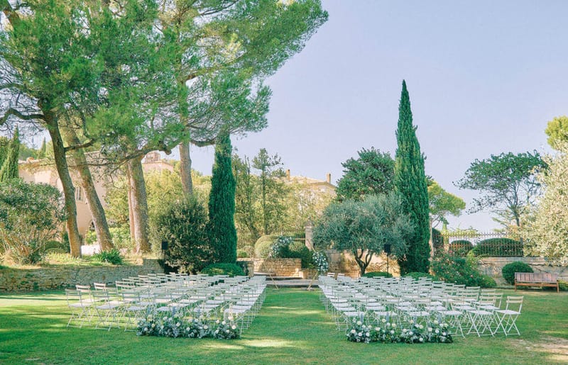Outdoor ceremony setup with white chairs and blue floral aisle markers on lawn before stone chateau