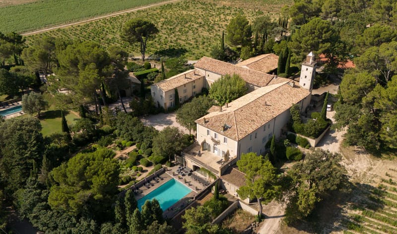 Aerial drone shot of a large Provençal château and its grounds, showing warm terracotta-tiled roofs, cream stone facades, and a distinctive tower with a pointed finial. The property features two visible swimming pools — one turquoise pool with a surrounding terrace and lounge chairs directly behind the main building, and a second pool visible to the upper left — along with manicured hedgerows, topiary, and a graveled central courtyard. The estate is bordered by vineyards in the upper portion of the frame and surrounded by mature pine and cypress trees. No people are visible in the shot. Potential venue feature image.