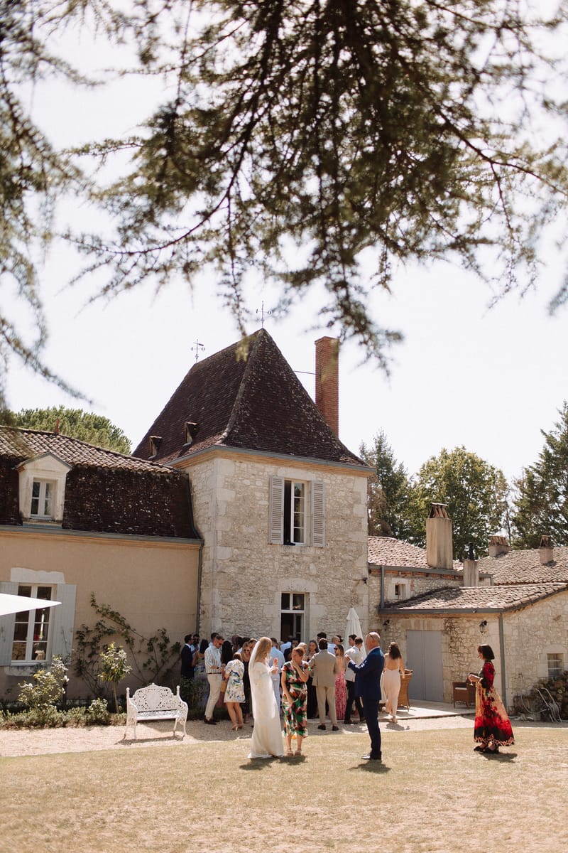 An outdoor cocktail hour taking place in the courtyard of a French stone manor house featuring a distinctive pointed tower with a dark slate roof, white shuttered windows, and terracotta tile roofing on the adjoining wings. Approximately 20–25 guests are mingling in small groups across the courtyard, with the bride visible in a white floor-length dress near the center foreground, accompanied by a groom in a navy suit. Guests are dressed in a mix of colorful summer attire including a bold green floral dress and a red patterned maxi dress. A white ornate garden bench is positioned to the left side of the courtyard. The shot is a wide, slightly elevated perspective framed by overhanging tree branches in the upper portion of the frame, captured in bright midday light. Potential venue feature image.