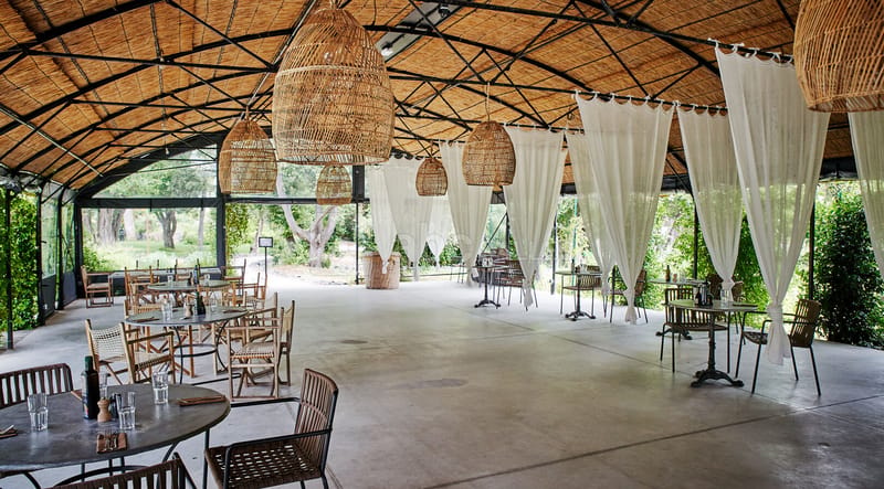 Thatched-roof open pavilion with wicker pendants, sheer curtains, and bistro chairs on polished concrete
